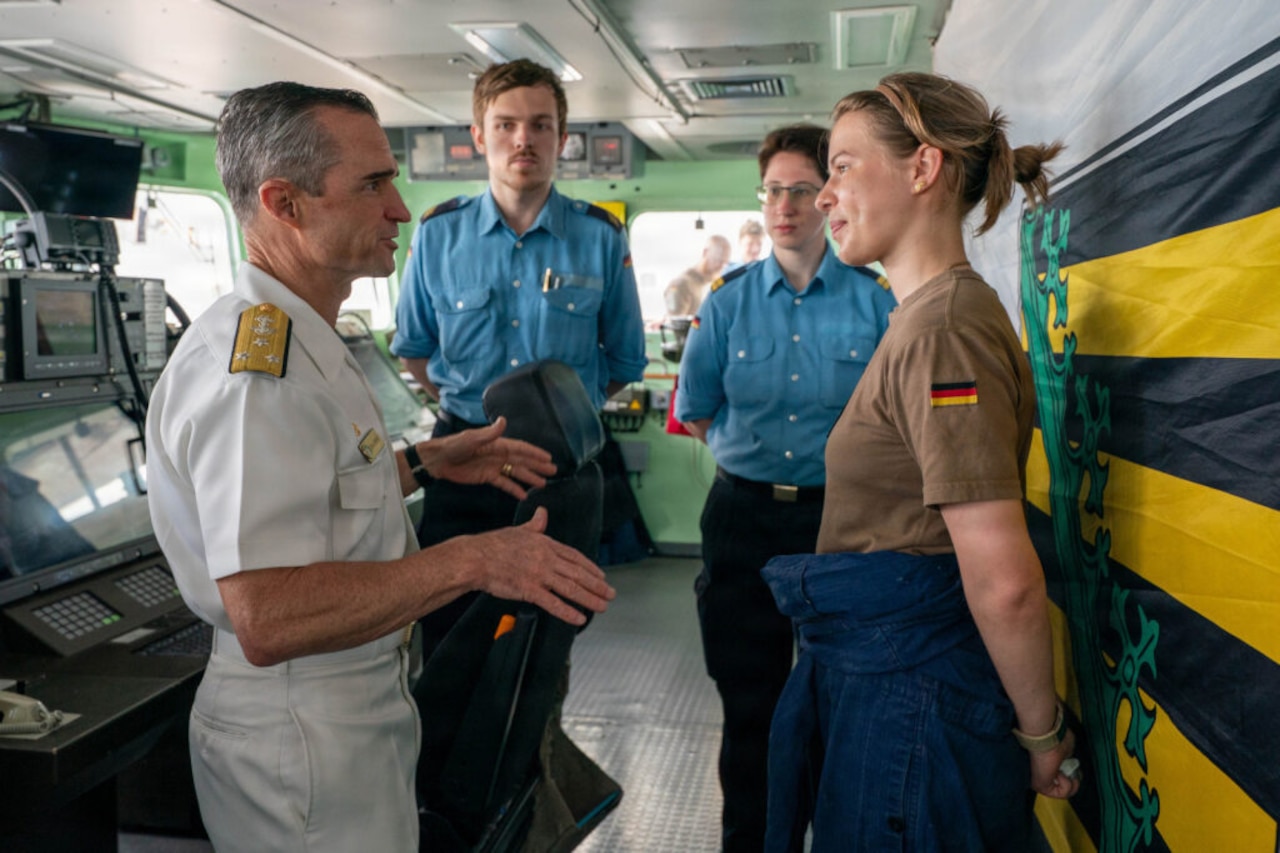 A man wearing a white military dress uniform talks to three other people in foreign military uniforms inside the bridge of a military ship.