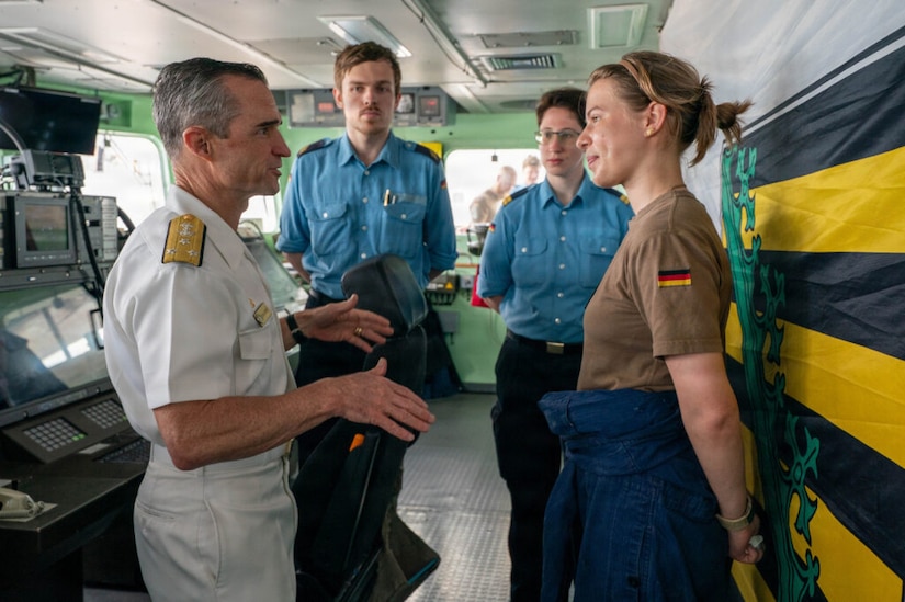 A man wearing a white military dress uniform talks to three other people in foreign military uniforms inside the bridge of a military ship.