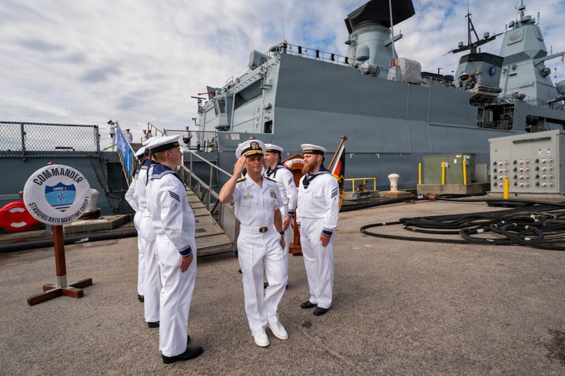Five men in military dress uniforms stand at attention as another man in similar attire standing between them renders a salute; a military ship is in the background.