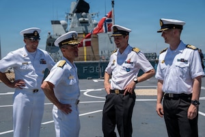Four men in military dress uniforms stand and chat on the deck of a military ship; the Norwegian flag is flying in the background.