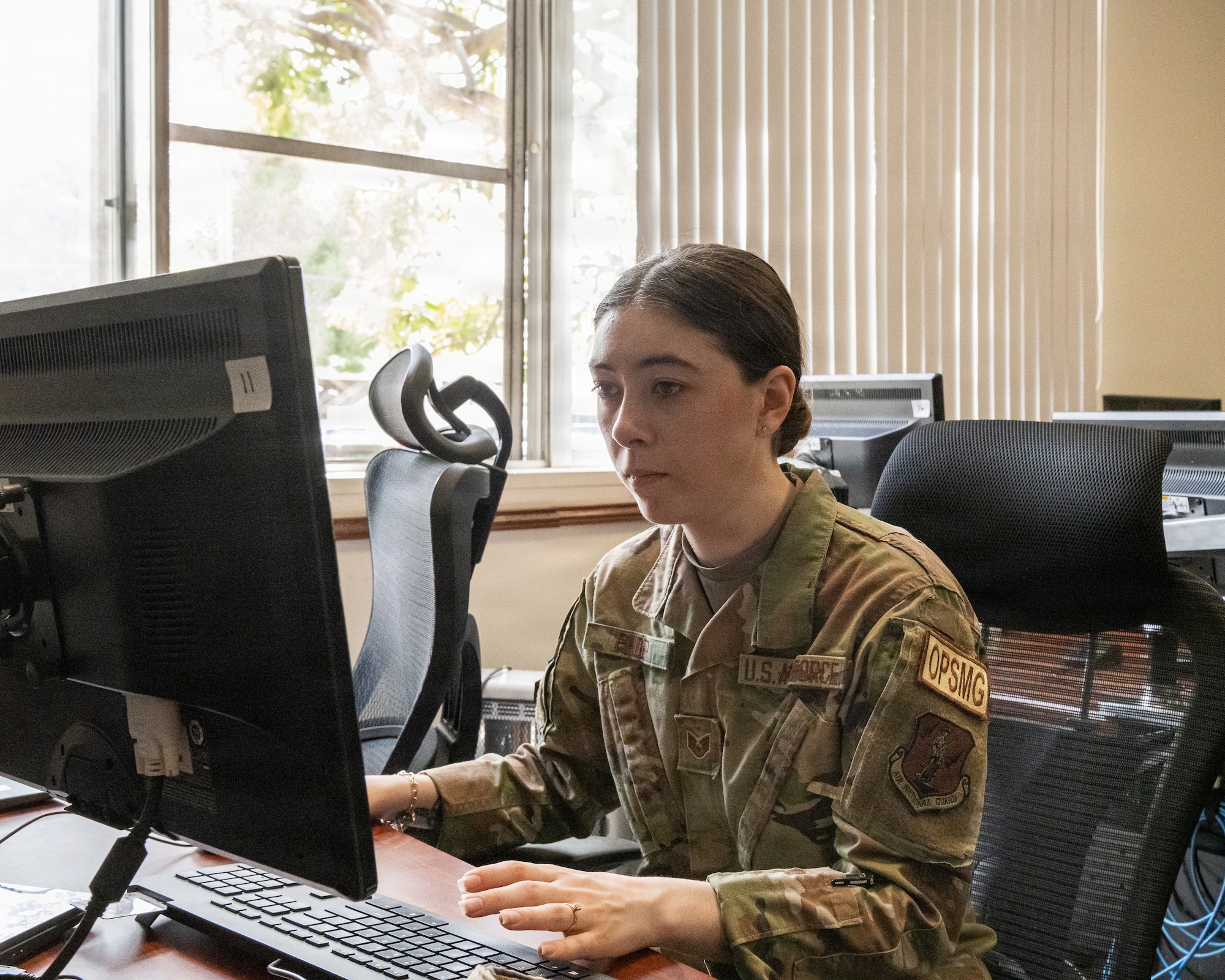 “Service member in camouflage uniform seated at a desk, typing on a computer in a sunlit office with multiple monitors and chairs visible in the background.