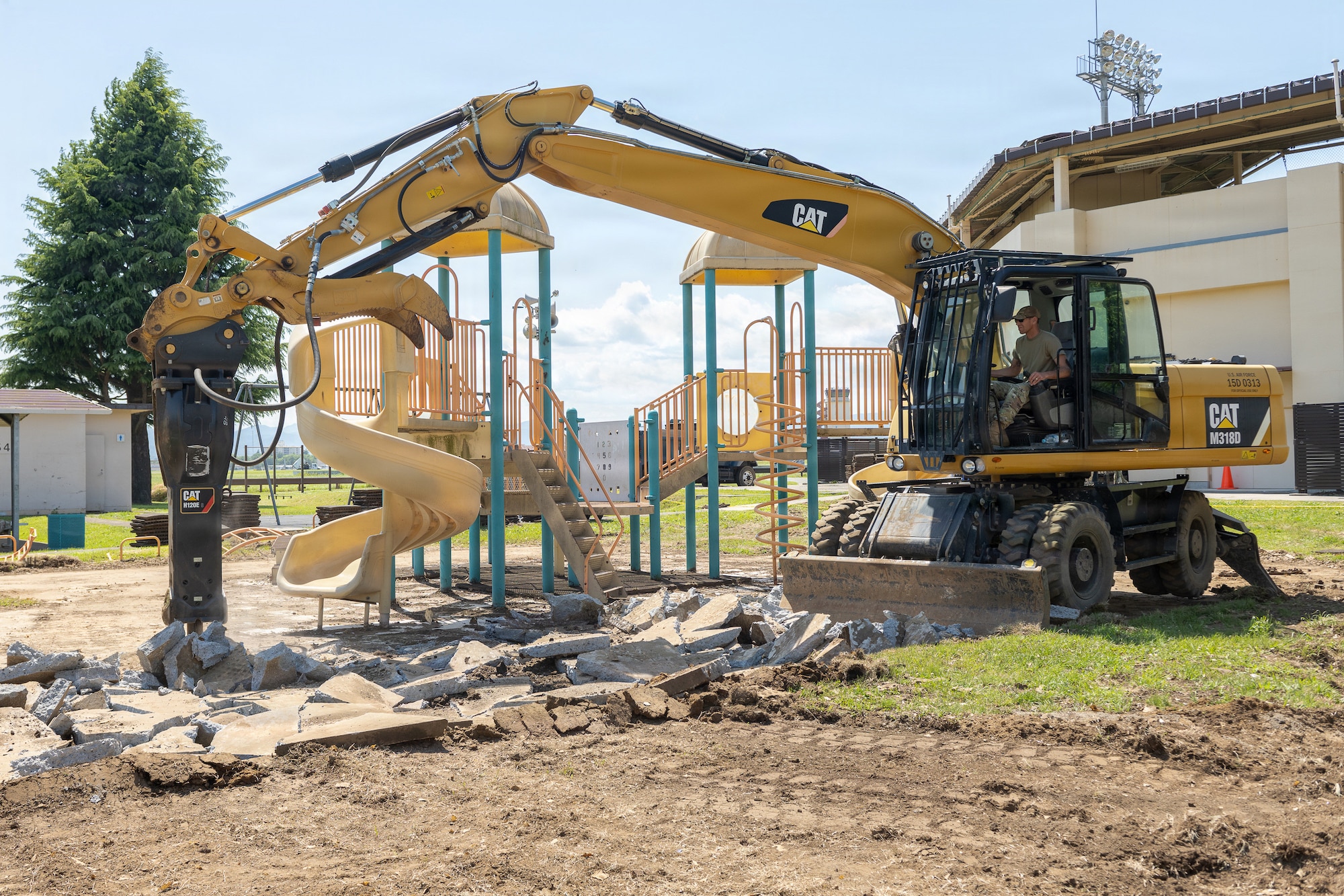 Yellow excavator breaking up dirt and debris beside a playground with a spiral slide, with a building and stadium lights in the background.