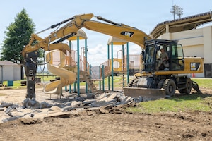 Yellow excavator breaking up dirt and debris beside a playground with a spiral slide, with a building and stadium lights in the background.
