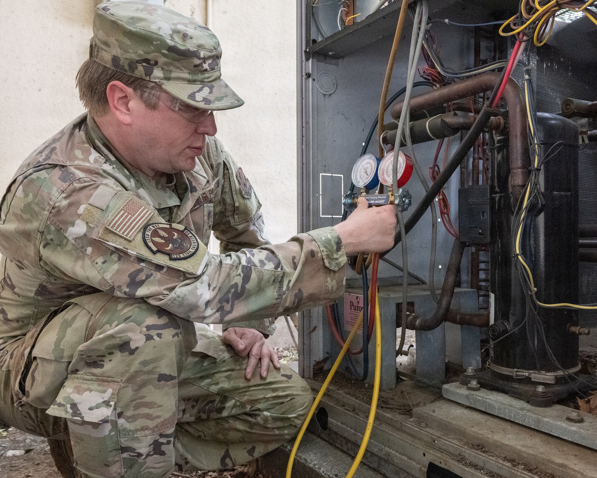 Service member in camouflage uniform checking an HVAC unit using a set of red, blue, and yellow pressure gauges connected to copper lines and wiring.