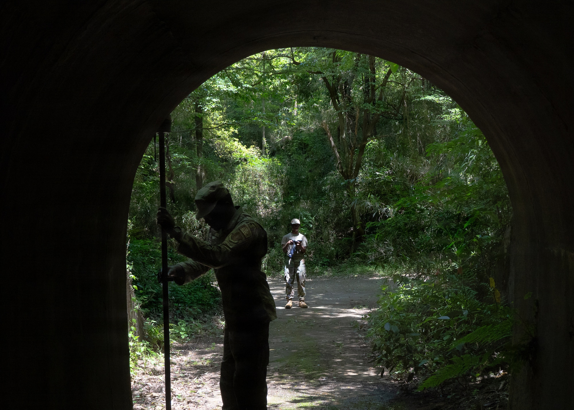 Two people in a forested area viewed from inside a dark tunnel; one stands at the entrance holding a long pole while another is farther down the sunlit path surrounded by dense greenery.
