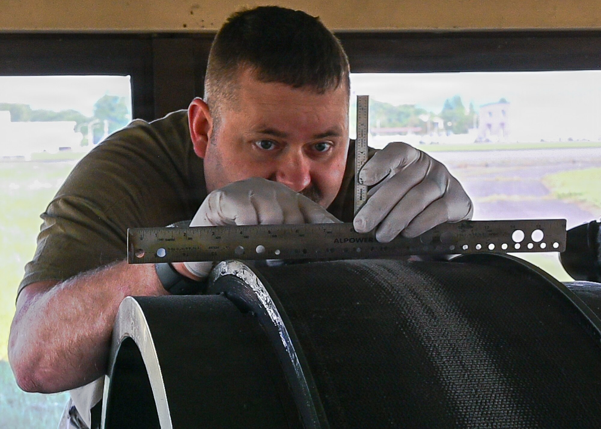 Person wearing white gloves using a metal ruler to measure or align a large cylindrical object in a workshop setting