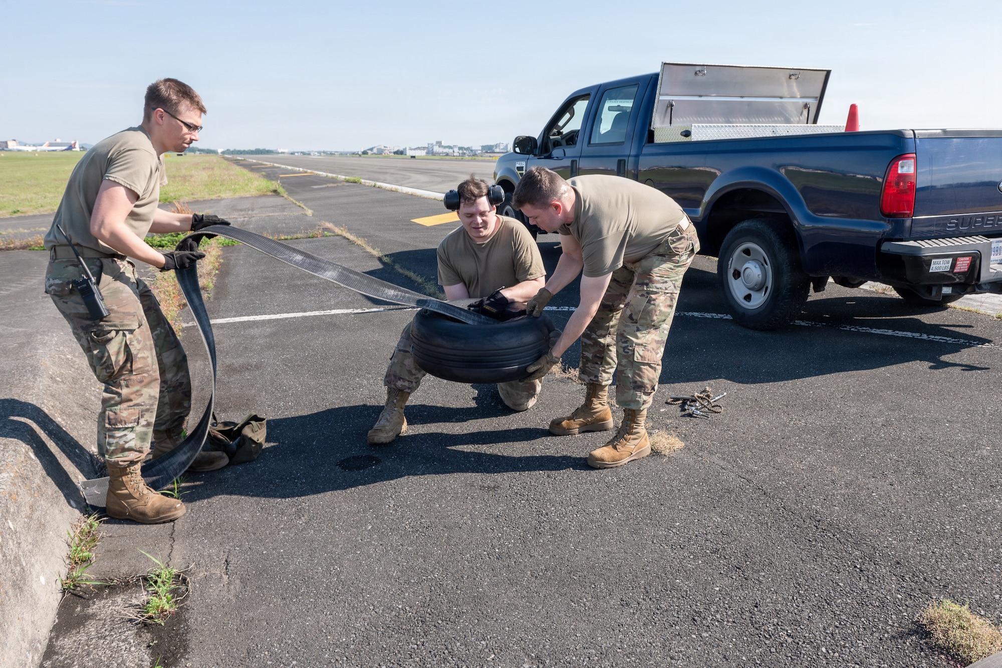 Three people in tan shirts and camouflage pants work together outdoors, handling a large black rubber object on a paved surface beside a blue pickup truck, with grassy fields and distant buildings in the background.