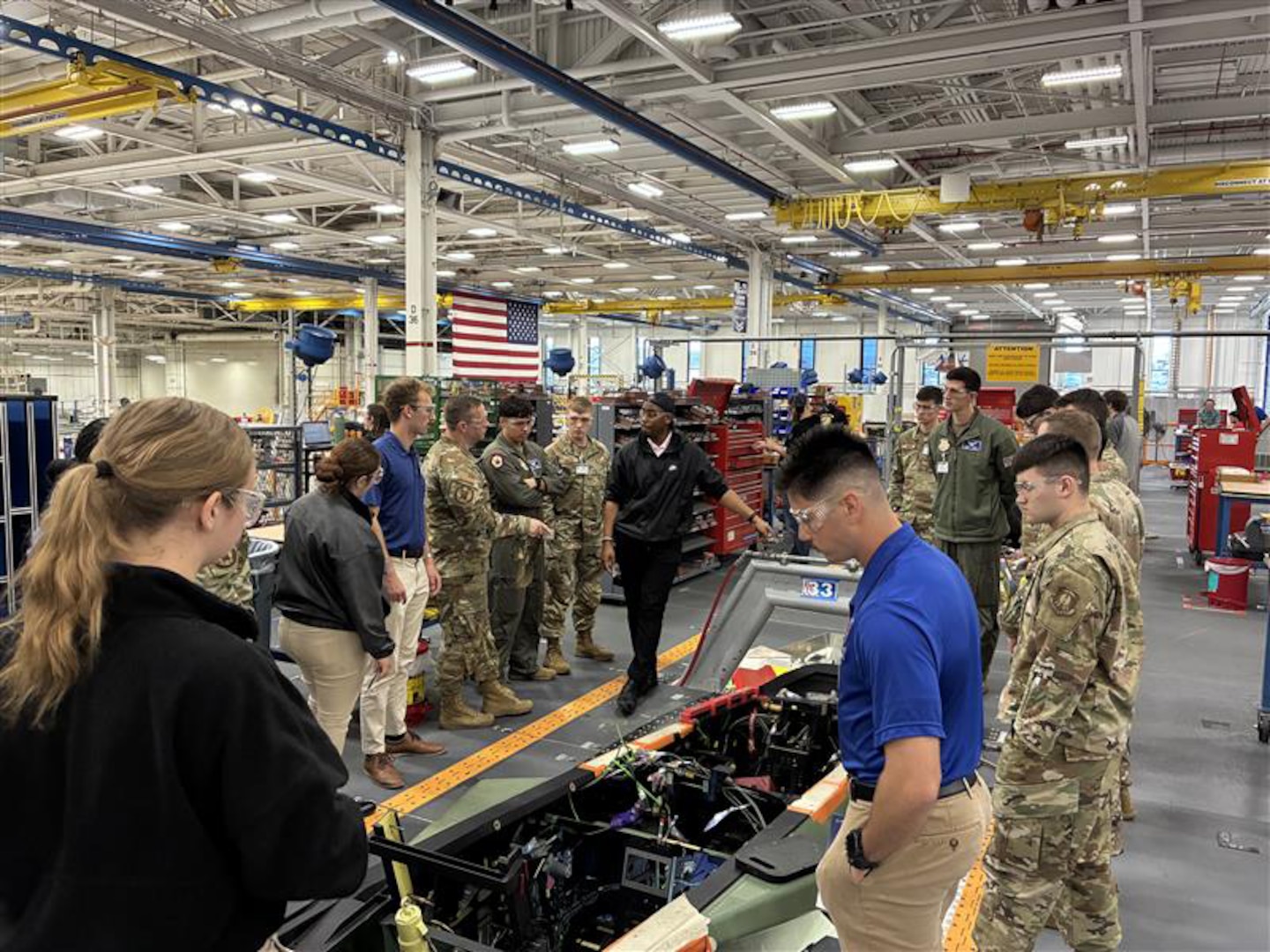 A group of men and women in military and civilian attire look at an aircraft part