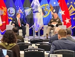 Two men sit in chairs on a stage. Military flags are in the background.