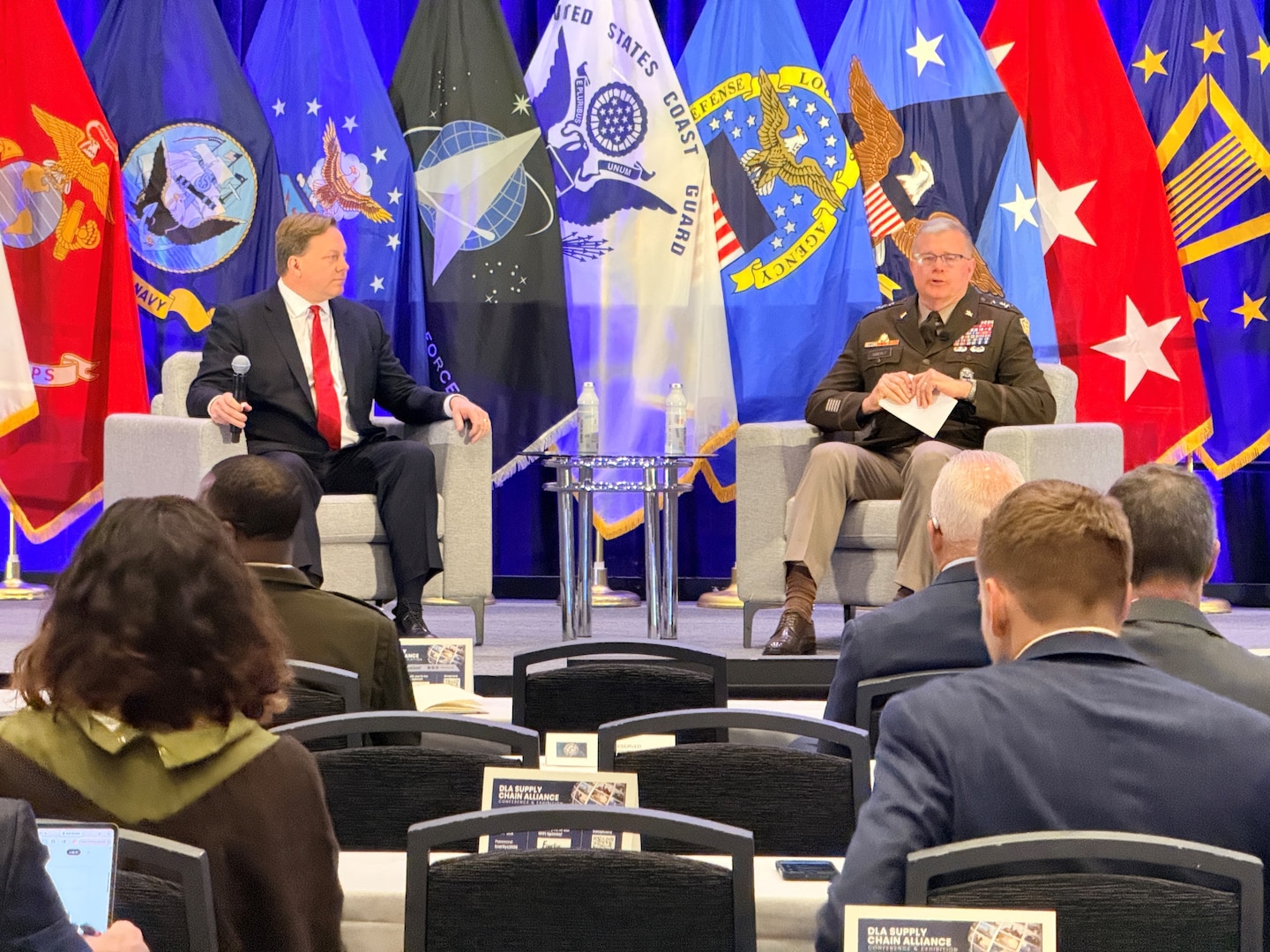 Two men sit in chairs on a stage. Military flags are in the background.
