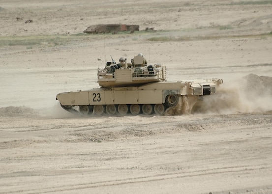 A side angle view of the M1 Abrams Tank rolling in the desert on a dirt road with one service member sitting on top. 