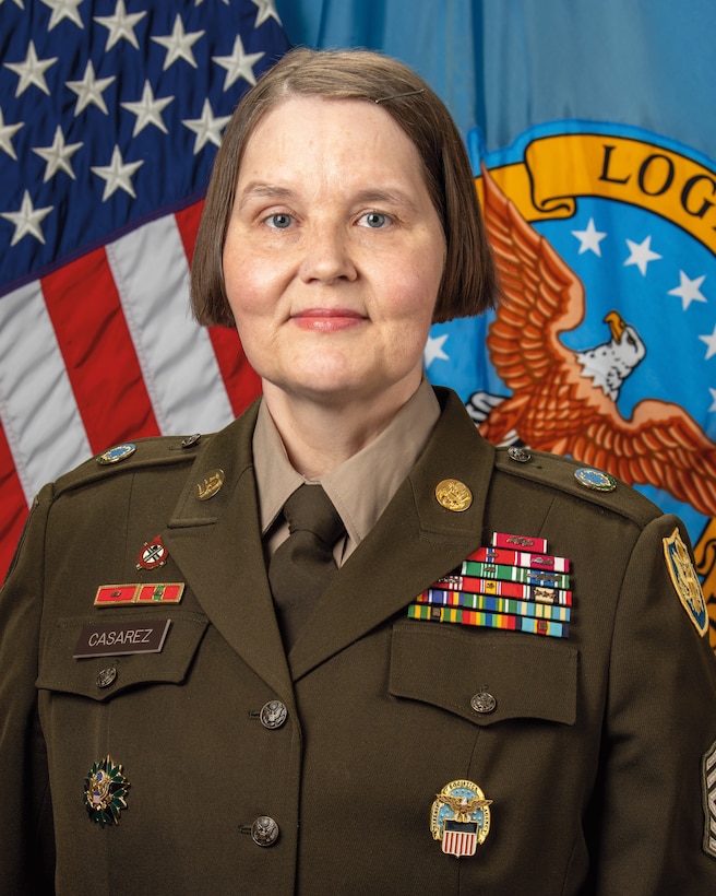 A headshot of a woman wearing an Army dress uniform. An American flag and the Army flag are in the background.