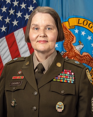 A headshot of a woman wearing an Army dress uniform. An American flag and the Army flag are in the background.