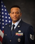 An Air Force Airman in dress uniform poses in front of an American flag.