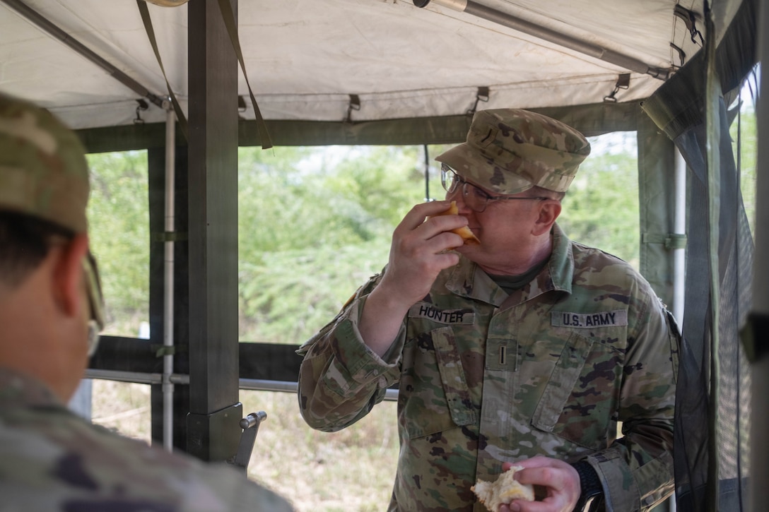 A soldier in a camouflage uniform and cap stands inside a screened tent, eating a meal. Another person is partially visible in the foreground.