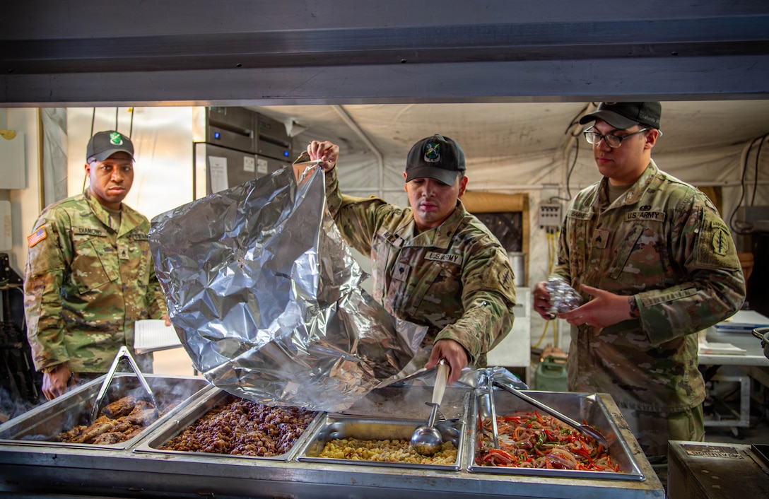 Three soldiers in camouflage uniforms are in a food service line. The soldier in the center is opening a large foil bag over a steam table that contains various hot food dishes. The soldier on the left is looking on, and the soldier on the right is holding a foil-wrapped item.