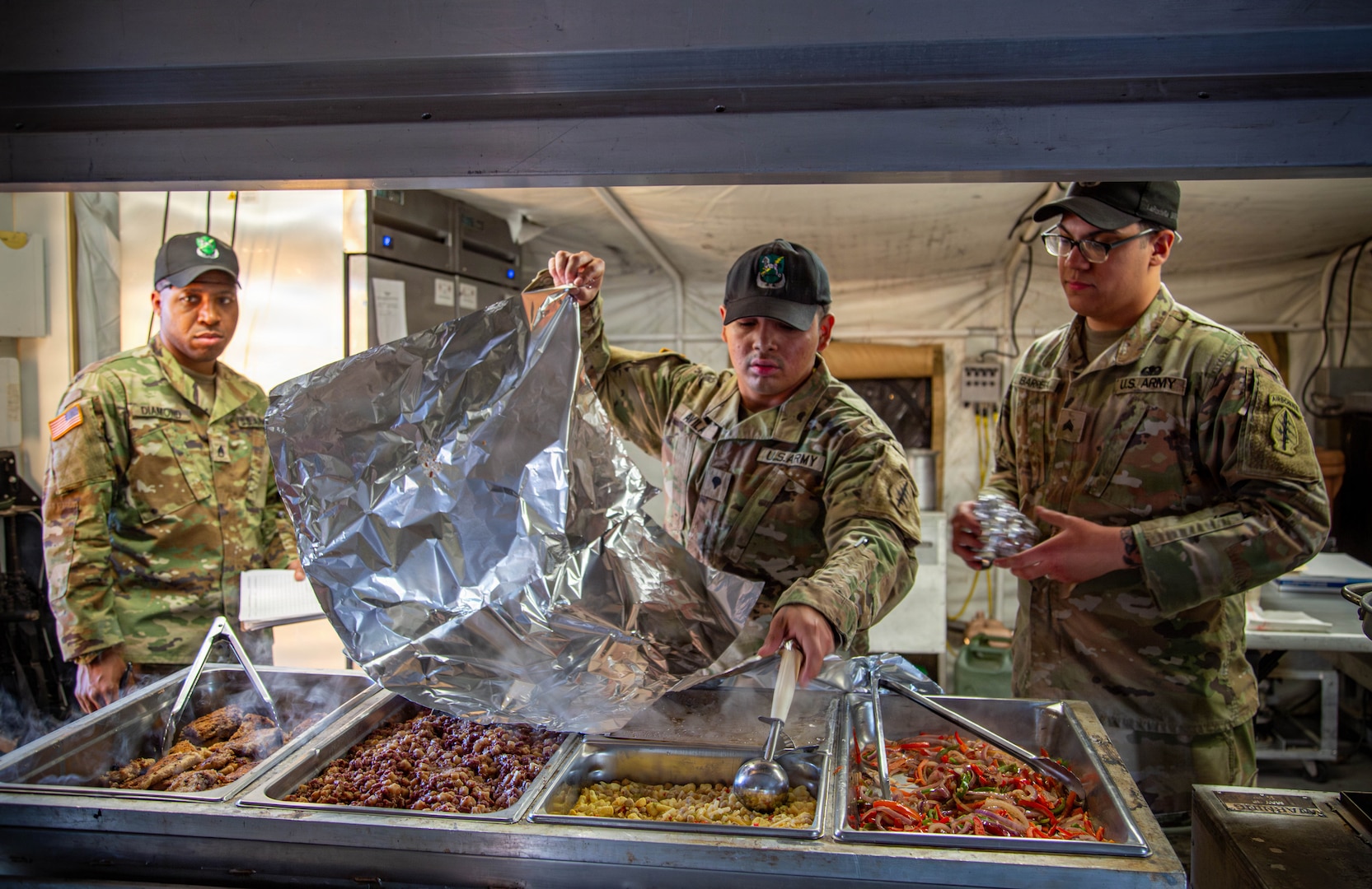 Three soldiers in camouflage uniforms are in a food service line. The soldier in the center is opening a large foil bag over a steam table that contains various hot food dishes. The soldier on the left is looking on, and the soldier on the right is holding a foil-wrapped item.