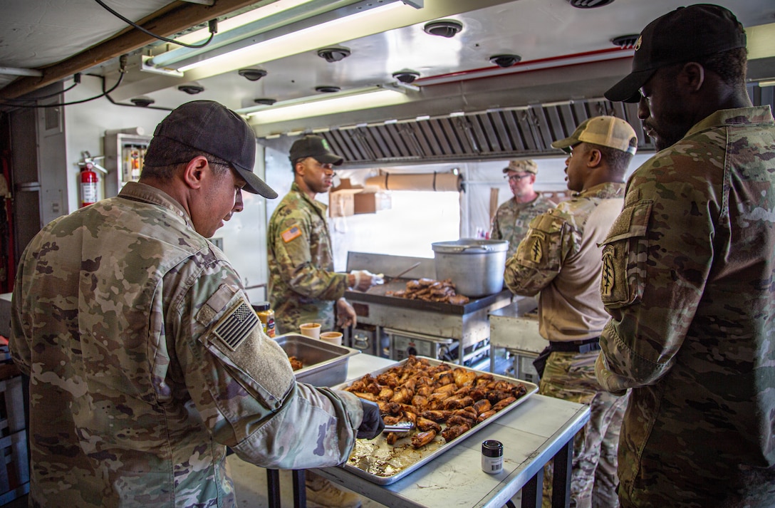Several soldiers in camouflage uniforms prepare food in a field kitchen. One soldier in the foreground is arranging pieces of fried chicken in a large serving pan. In the background, other soldiers are cooking and serving food.