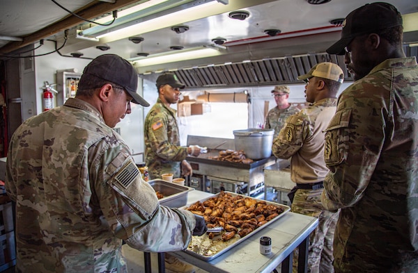 Several soldiers in camouflage uniforms prepare food in a field kitchen. One soldier in the foreground is arranging pieces of fried chicken in a large serving pan. In the background, other soldiers are cooking and serving food.