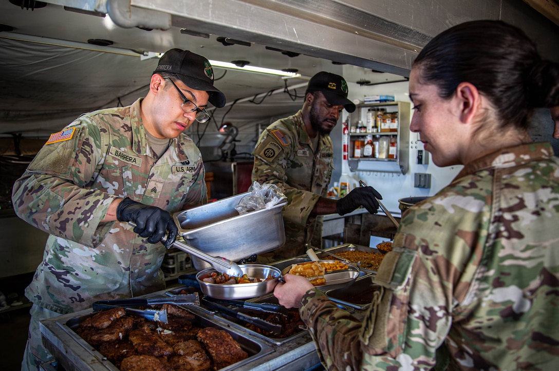 Two soldiers in camouflage uniforms serve a hot meal to a fellow soldier from a field kitchen food line.