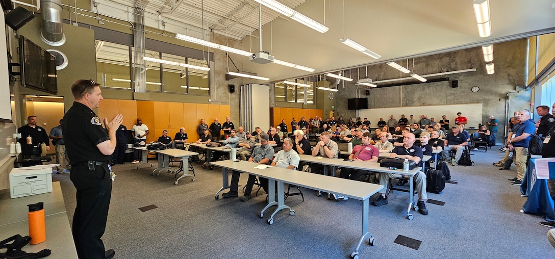 A police officer in a dark uniform stands at the front of a classroom, addressing a large, seated audience during what appears to be a training session or briefing. The room is filled with people listening attentively from their seats at long tables.