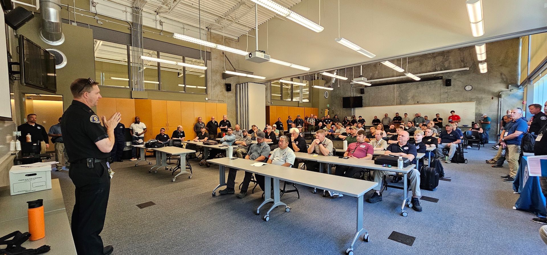 A police officer in a dark uniform stands at the front of a classroom, addressing a large, seated audience during what appears to be a training session or briefing. The room is filled with people listening attentively from their seats at long tables.