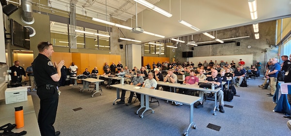 A police officer in a dark uniform stands at the front of a classroom, addressing a large, seated audience during what appears to be a training session or briefing. The room is filled with people listening attentively from their seats at long tables.