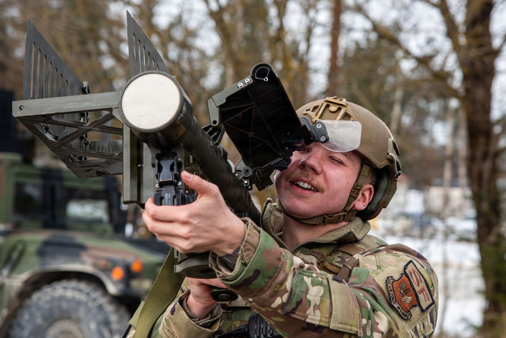 A soldier in a helmet and camouflage uniform is aiming a shoulder-fired missile launcher. He is looking through the weapon's sight with a focused expression. A military vehicle is partially visible in the background.