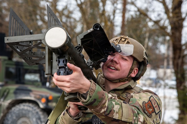 A soldier in a helmet and camouflage uniform is aiming a shoulder-fired missile launcher. He is looking through the weapon's sight with a focused expression. A military vehicle is partially visible in the background.