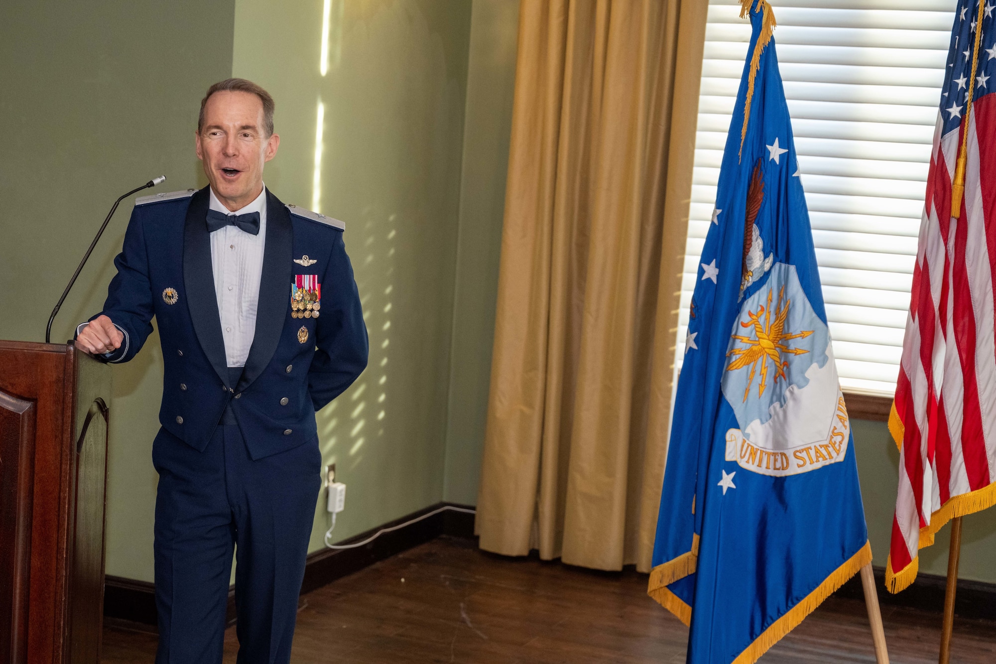 A man delivers a speech wearing his mess dress uniform while standing next to the Air Force flag and the U.S. flag.