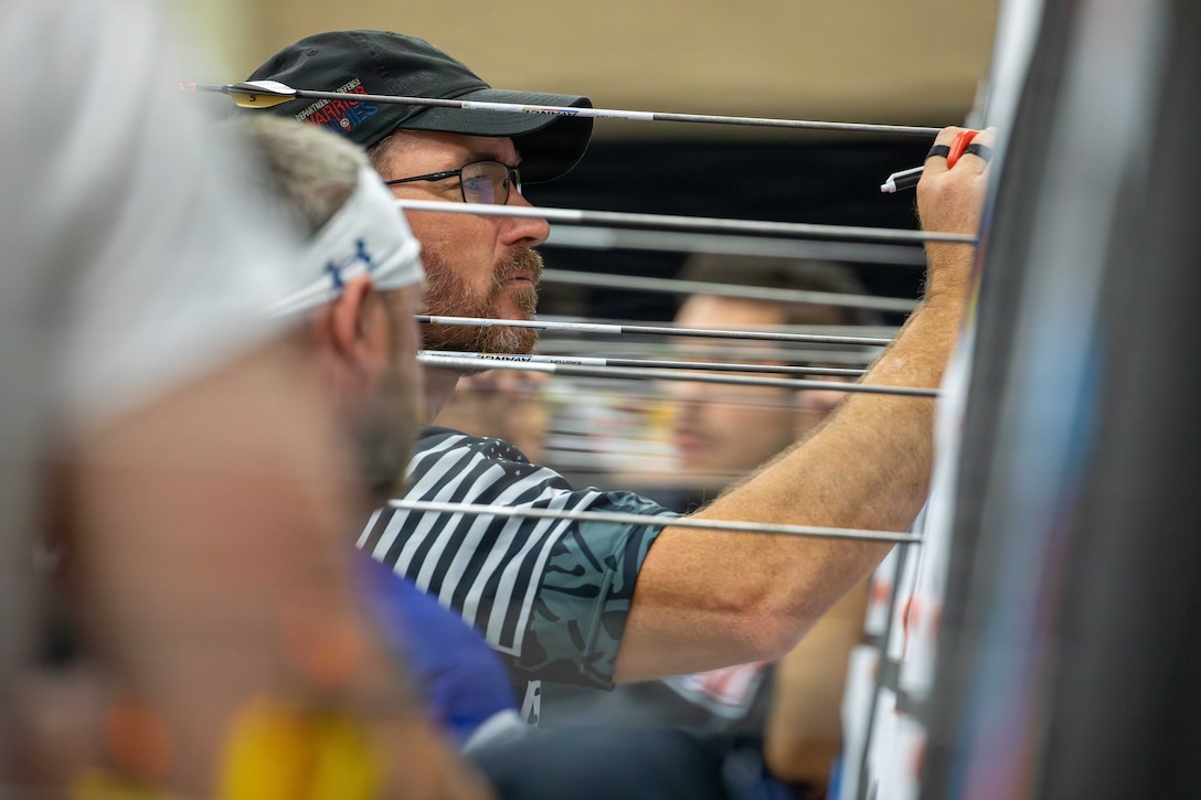 A man in athletic clothing uses a pen to make a mark on a board that has arrows sticking out of it as other athletes stand near him.