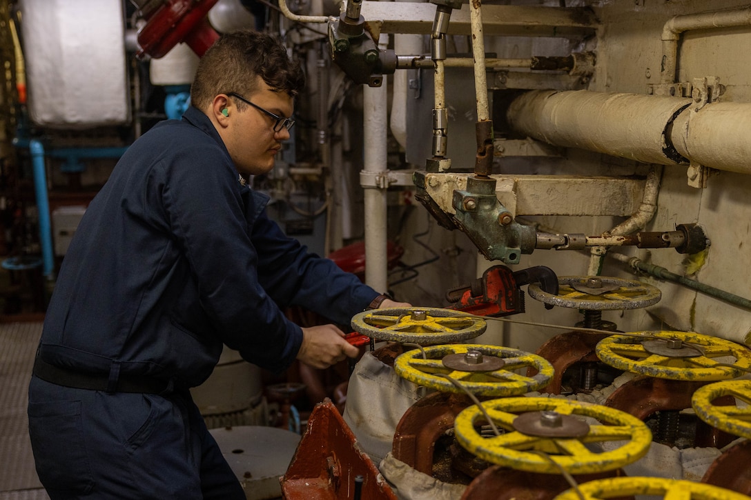 A sailor in a blue uniform and glasses is in the engine room of a ship, turning one of many large, yellow-handled valves.