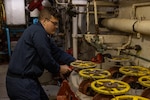 A sailor in a blue uniform and glasses is in the engine room of a ship, turning one of many large, yellow-handled valves.