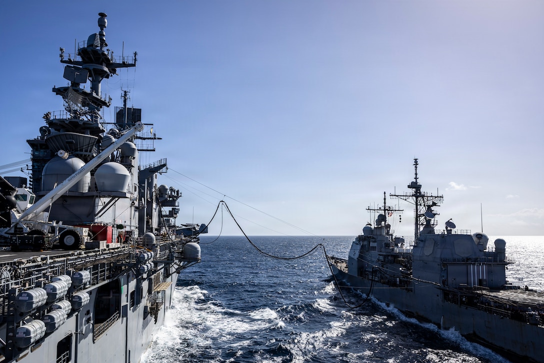 Two large naval warships are sailing side-by-side on the open ocean under a clear blue sky. A thick hose connects the two ships, indicating a refueling-at-sea operation is in progress.