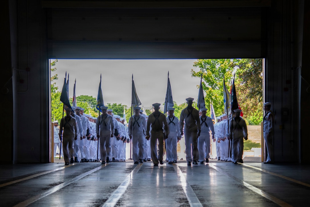Sailors march into a hall from the outside in formation, with some carrying flags.