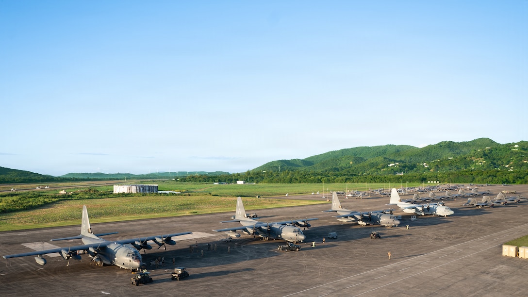 A line of large, gray military transport aircraft is parked on an airfield tarmac. Green, rolling hills are visible in the background under a clear sky.