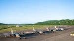 A line of large, gray military transport aircraft is parked on an airfield tarmac. Green, rolling hills are visible in the background under a clear sky.