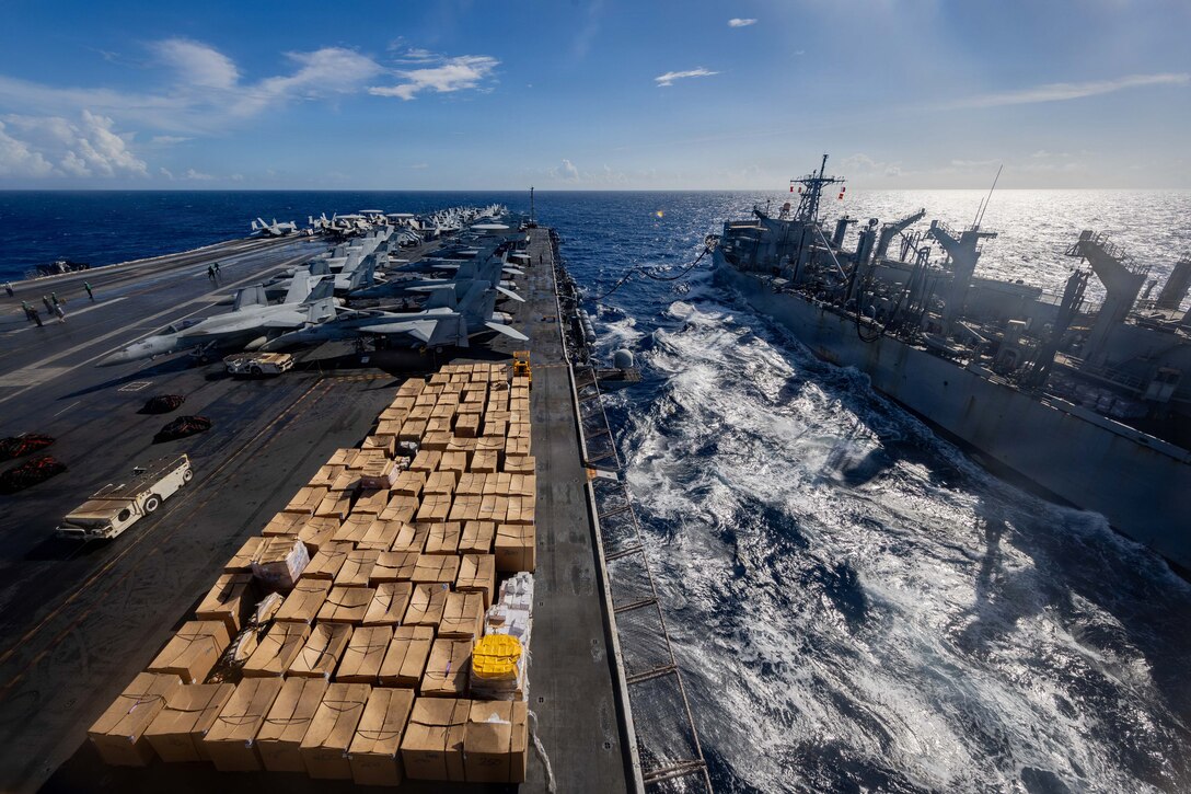 From a high vantage point on an aircraft carrier, the flight deck is seen covered with neatly stacked cardboard boxes during a replenishment operation. A second naval vessel sails alongside on the choppy open sea under a partly cloudy sky.