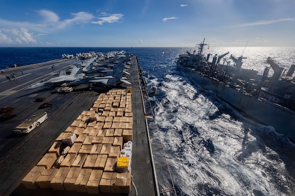 From a high vantage point on an aircraft carrier, the flight deck is seen covered with neatly stacked cardboard boxes during a replenishment operation. A second naval vessel sails alongside on the choppy open sea under a partly cloudy sky.