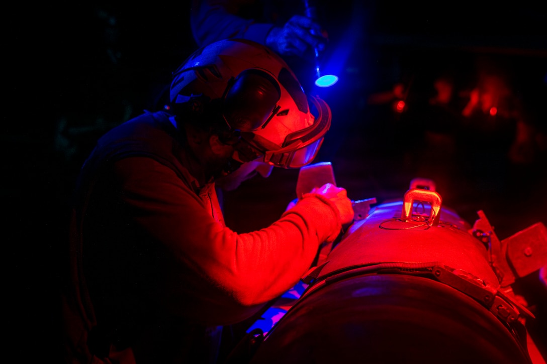 Red and blue light is reflected on a sailor and part of the equipment they are working on at night. Another sailor holds a flashlight.