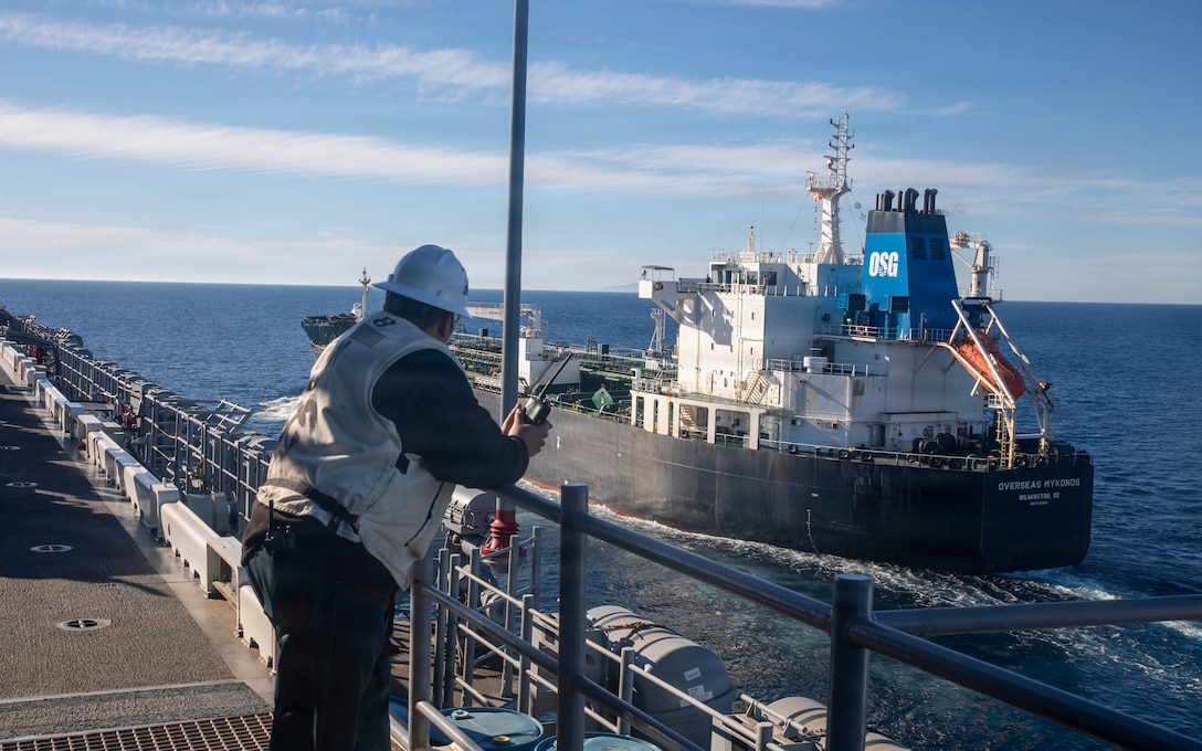 From the deck of a ship, a person wearing a white hard hat and vest watches a large black and white tanker ship as it sails alongside on the open sea.