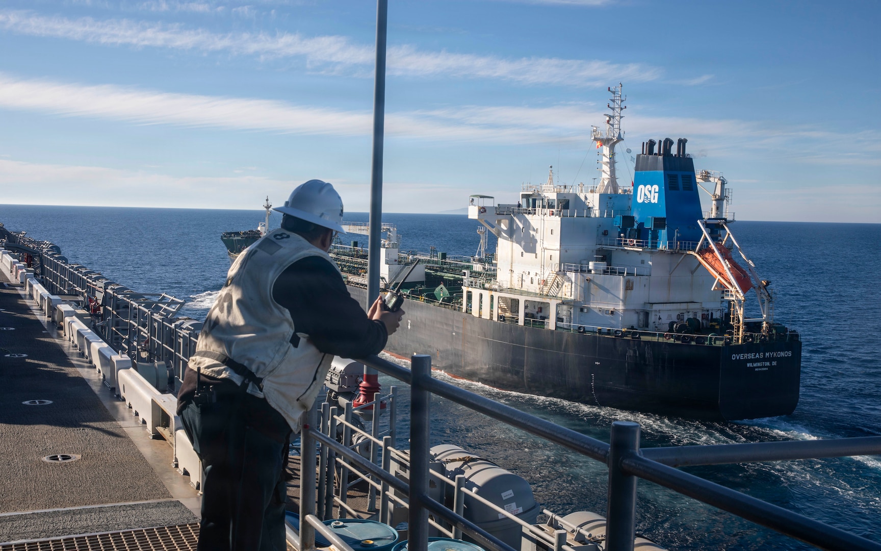 From the deck of a ship, a person wearing a white hard hat and vest watches a large black and white tanker ship as it sails alongside on the open sea.