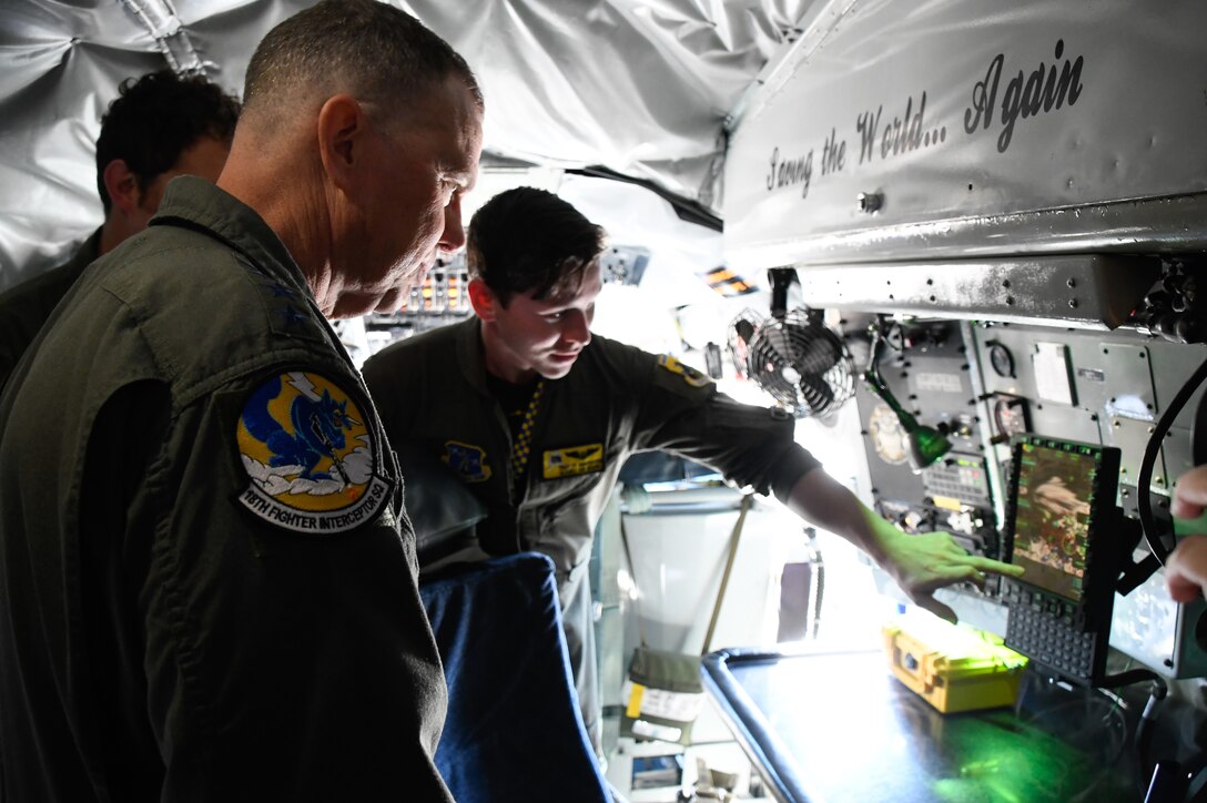 Inside a military aircraft, a uniformed airman points to a laptop screen as he briefs a senior officer.