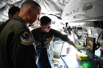 Inside a military aircraft, a uniformed airman points to a laptop screen as he briefs a senior officer.