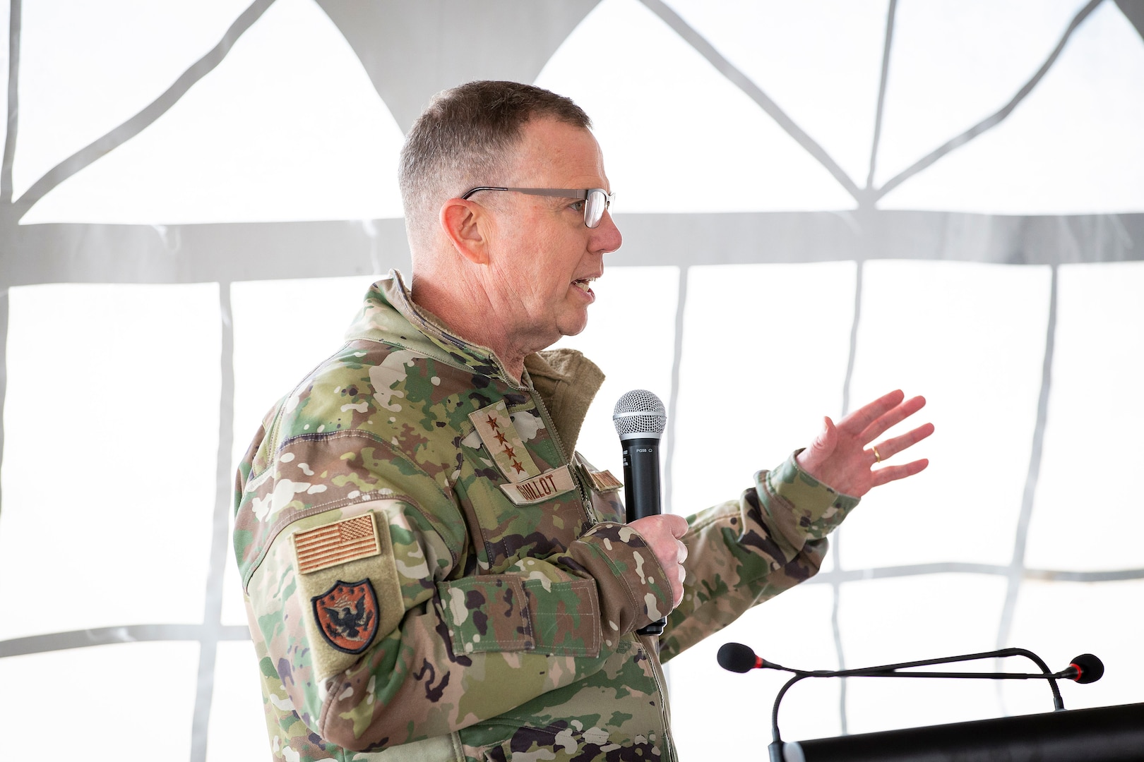 A four-star Air Force general holds a microphone and speaks to a crowd from a podium.