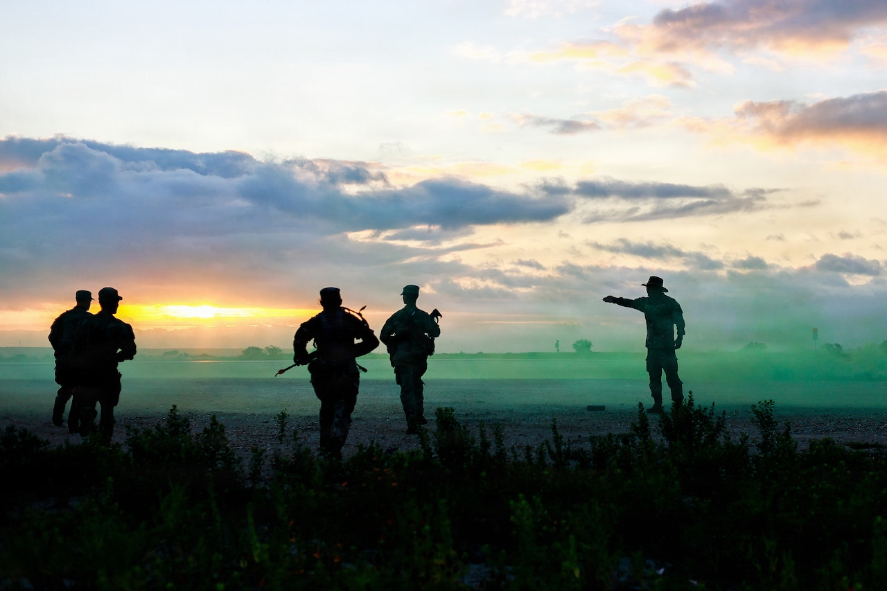 A service member wearing a cavalry hat and military battle dress uniform is seen in silhouette pointing with his arm as four other silhouetted service members move in the direction he is pointing while wearing battle dress uniforms and patrol caps, while carrying rifles in a field as the sun is low on the horizon.