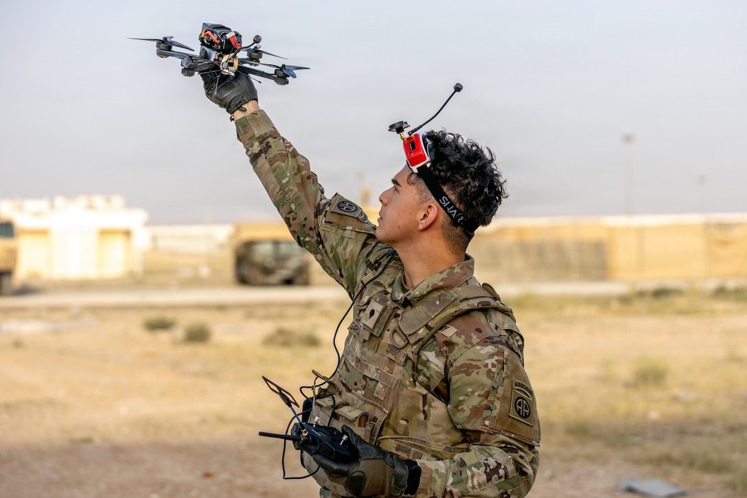 A soldier wearing a camouflage uniform and electronic goggles on his forehead holds up a small aerial drone with his right hand while holding a controller in his left hand in a desert environment, with buildings in the background.