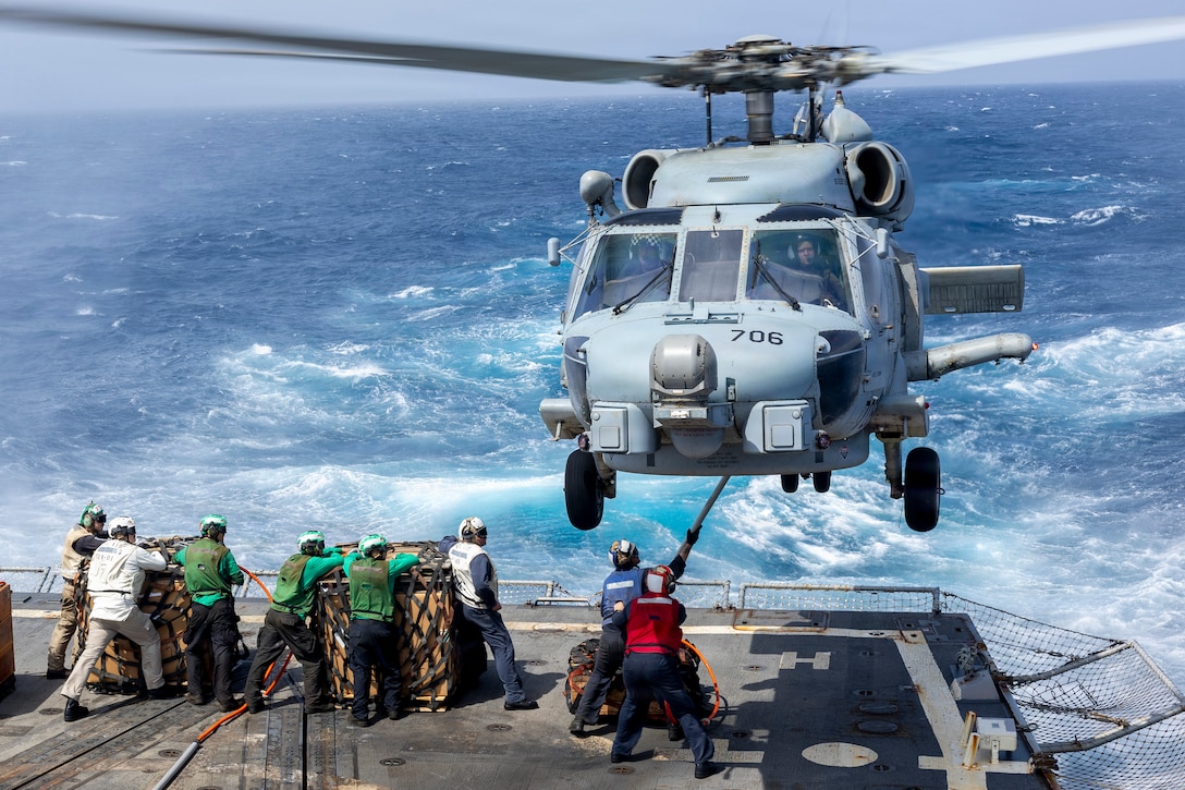 Two sailors on the deck of a ship at sea attach a cargo pallet to a hovering military helicopter while six other sailors wait nearby with two other pallets. The sailors are wearing white, green, red or blue vests.