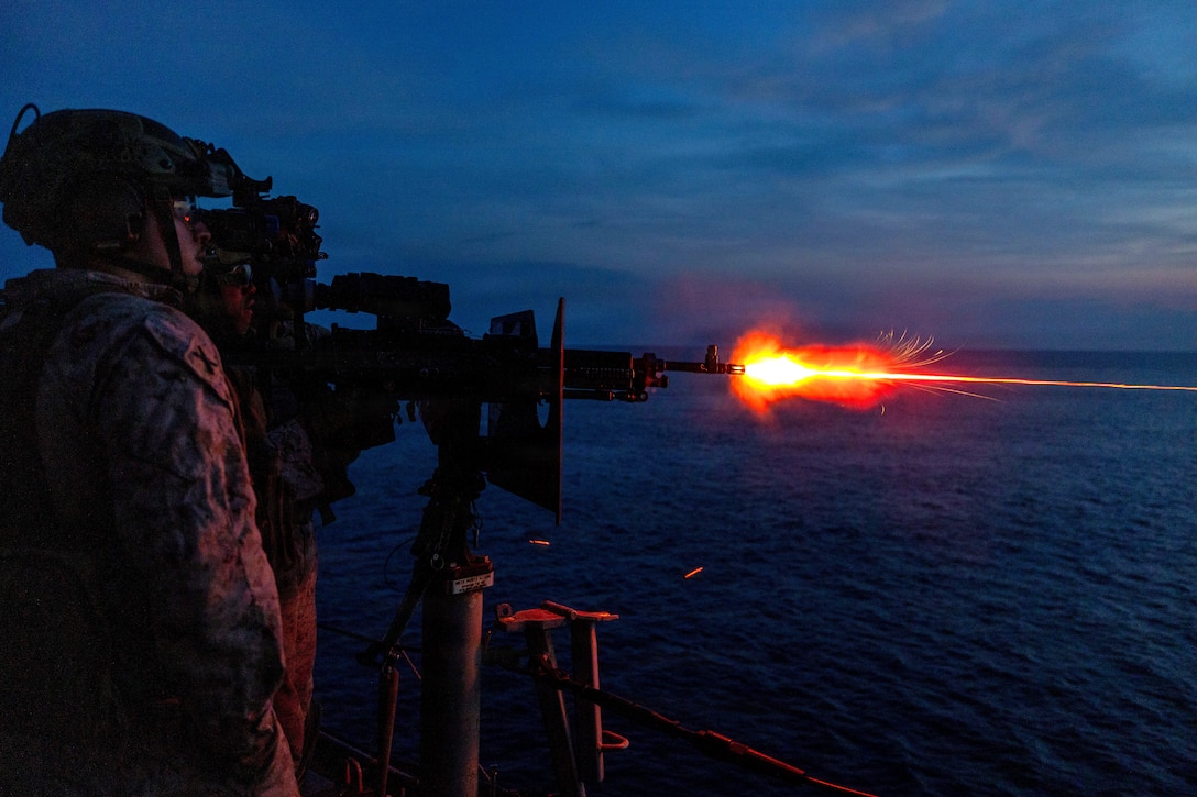 Flames blaze from the muzzle as tracer rounds are fired from a machine gun by a Marine wearing night-vision equipment, as another Marine, also with night-vision equipment, stands nearby in dark conditions on a ship, with an ocean in the background.