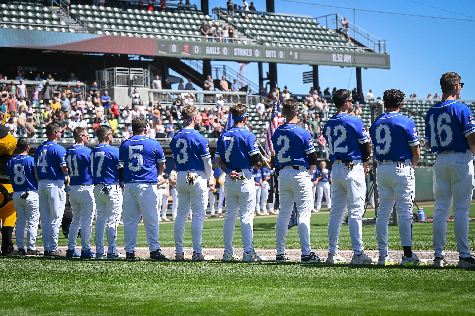 A long line of Hill Air Force Base baseball players stands shoulder-to-shoulder along the baseline of a professional stadium field, holding their baseball caps over their hearts during the playing of the national anthem.